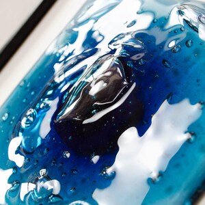 May include: A square glass art piece featuring a layered design of deep navy blue and vibrant turquoise tones. The surface has a glossy, melted appearance with numerous small air bubbles trapped within the glass, creating a textured, liquid effect.