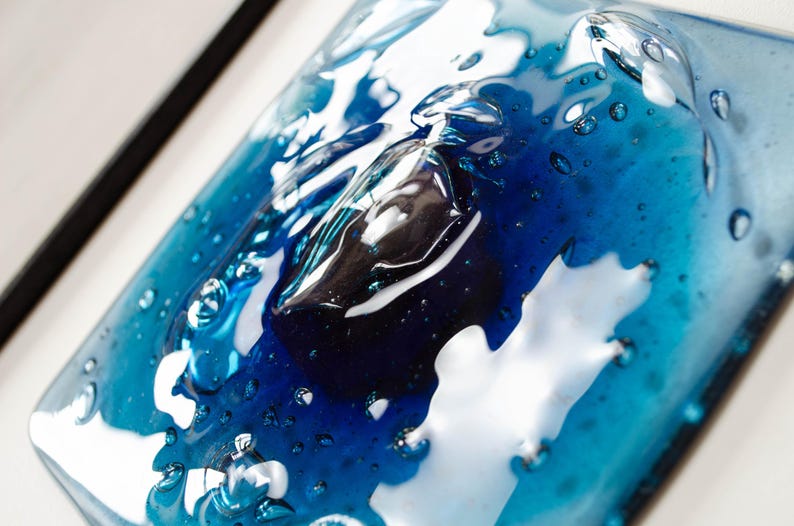 May include: A square glass art piece featuring a layered design of deep navy blue and vibrant turquoise tones. The surface has a glossy, melted appearance with numerous small air bubbles trapped within the glass, creating a textured, liquid effect.
