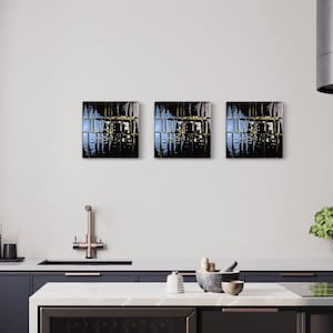 May include: Three square abstract art pieces with dark, reflective surfaces and gold accents are mounted on a white wall. The artwork is displayed above a kitchen counter with a sink, tap, and various kitchen accessories. The overall colour scheme is dark and modern.