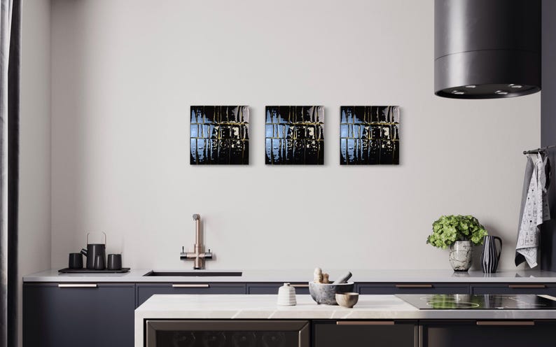 May include: Three square abstract art pieces with dark, reflective surfaces and gold accents are mounted on a white wall. The artwork is displayed above a kitchen counter with a sink, tap, and various kitchen accessories. The overall colour scheme is dark and modern.