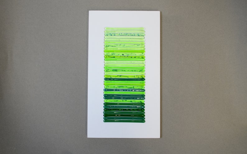 May include: A white rectangular frame with a vertical row of 25 green glass tubes, each with a different shade of green.