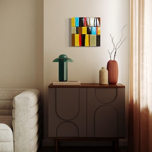 Könnte beinhalten: Ein modernes Wohnzimmer mit einer dunkelgrünen Tischlampe, einem grauen Schrank und einem farbenfrohen abstrakten Wandkunstwerk. Das Kunstwerk besteht aus rechteckigen Blöcken in verschiedenen Farben. Ein Sofa und dekorative Vasen vervollständigen den Raum.