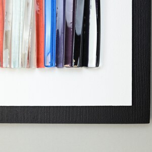 Pu&ograve; includere: Un'opera d'arte incorniciata che presenta un design astratto colorato realizzato con bacchette di vetro. Le bacchette sono disposte in uno schema verticale e sono di diverse lunghezze e colori, tra cui blu, rosso, nero, bianco e viola.