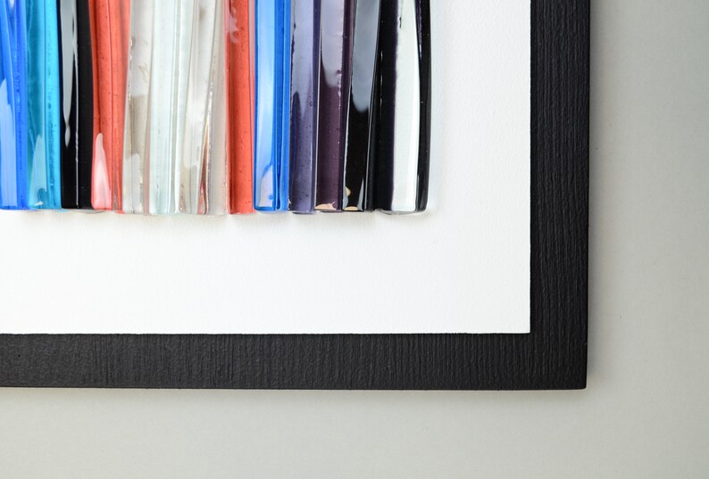 Pu&ograve; includere: Un'opera d'arte incorniciata che presenta un design astratto colorato realizzato con bacchette di vetro. Le bacchette sono disposte in uno schema verticale e sono di diverse lunghezze e colori, tra cui blu, rosso, nero, bianco e viola.