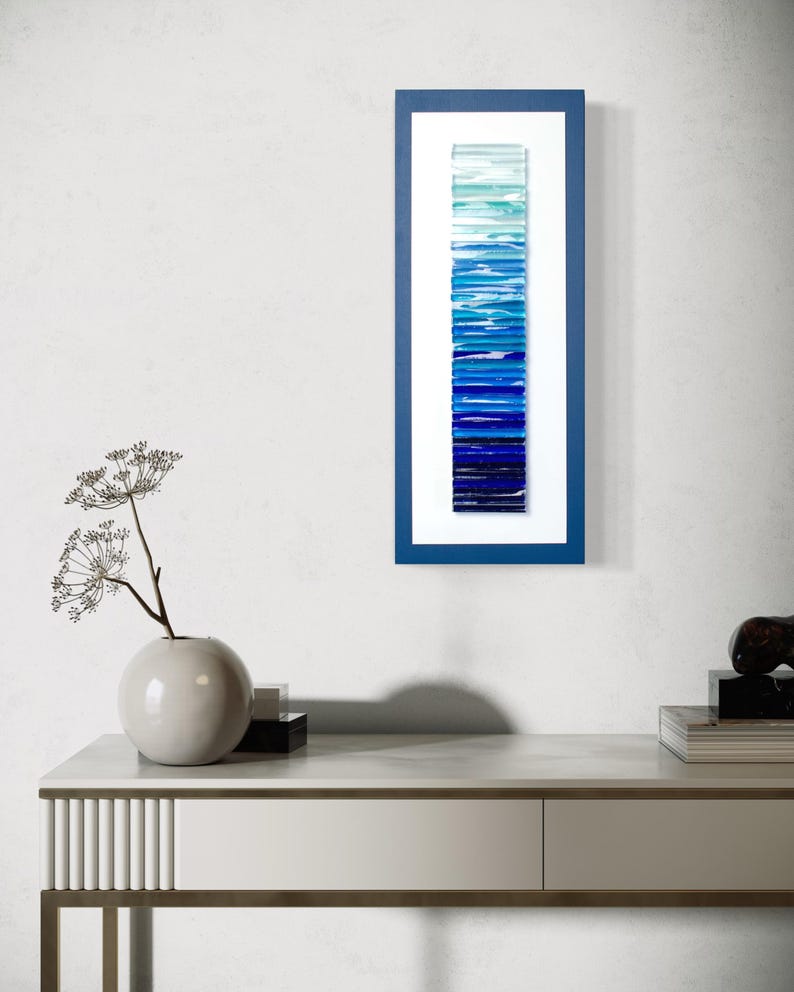 May include: A framed wall art piece with a gradient of blue glass tiles, ranging from light blue to dark blue, creating a vertical ombre effect.