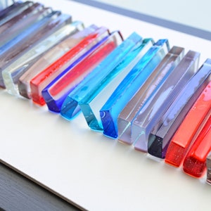 May include: A wall art piece made of glass rods in various colors, including blue, red, purple, and gray. The rods are arranged in a staggered pattern, creating a modern and abstract design.