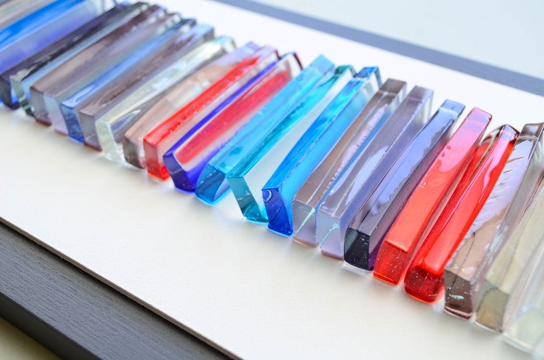 May include: A wall art piece made of glass rods in various colors, including blue, red, purple, and gray. The rods are arranged in a staggered pattern, creating a modern and abstract design.