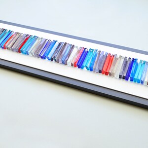 May include: A framed wall art piece featuring a long row of colorful glass rods in shades of blue, red, and gray. The rods are arranged in a staggered pattern, creating a dynamic and modern look.
