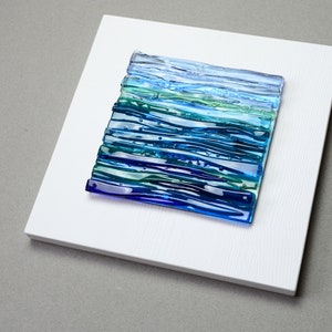 May include: A square, abstract glass art piece with a layered design of blues and greens, mounted on a white wooden panel. The glass art features horizontal lines and a textured appearance, evoking a sense of water or a landscape. The artwork is set against a neutral gray background.