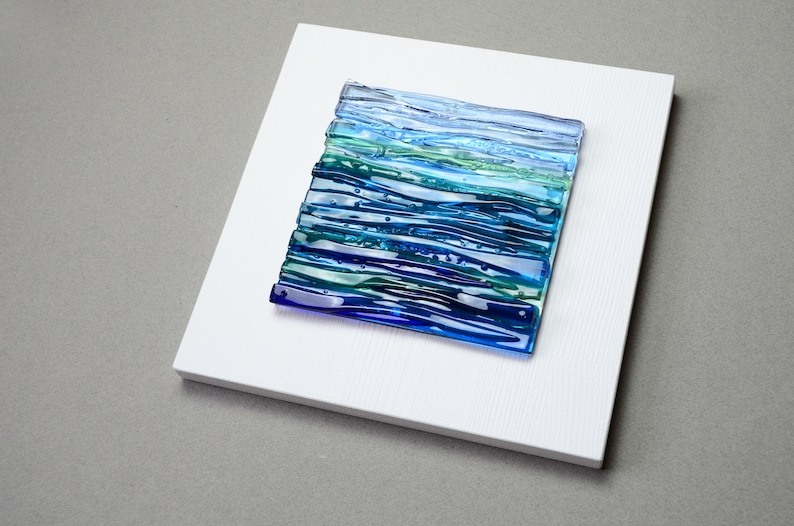 May include: A square, abstract glass art piece with a layered design of blues and greens, mounted on a white wooden panel. The glass art features horizontal lines and a textured appearance, evoking a sense of water or a landscape. The artwork is set against a neutral gray background.