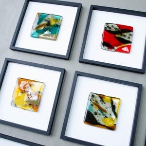May include: A collection of square wall art pieces featuring fused glass tiles in various colors including teal, red, yellow, and black. Each glass tile is mounted on a white background within a thin black square frame, arranged in a grid pattern.