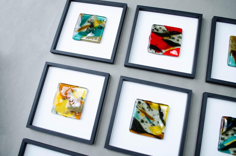 May include: A collection of square wall art pieces featuring fused glass tiles in various colors including teal, red, yellow, and black. Each glass tile is mounted on a white background within a thin black square frame, arranged in a grid pattern.