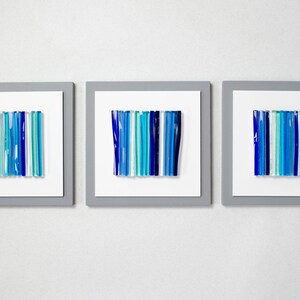 Pu&ograve; includere: Tre pezzi di arte da parete incorniciati con disegni astratti realizzati in vetro blu e turchese. I pezzi di vetro sono disposti in linee verticali.