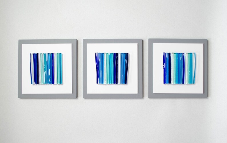 Pu&ograve; includere: Tre pezzi di arte da parete incorniciati con disegni astratti realizzati in vetro blu e turchese. I pezzi di vetro sono disposti in linee verticali.