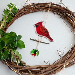 May include: A grapevine wreath with a red cardinal ornament perched on a branch. The cardinal is hanging from the wreath with a small holly ornament. Green ivy and fairy lights are also attached to the wreath.
