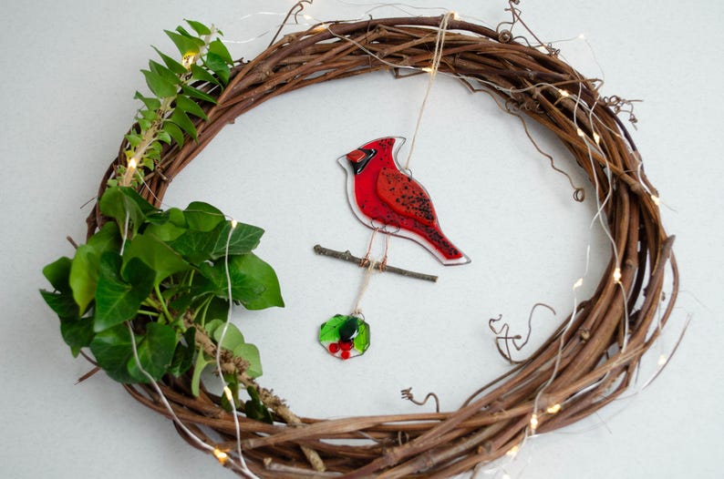 May include: A grapevine wreath with a red cardinal ornament perched on a branch. The cardinal is hanging from the wreath with a small holly ornament. Green ivy and fairy lights are also attached to the wreath.