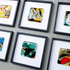 May include: Six square black frames containing individual fused glass art pieces. Each piece features a unique abstract composition of vibrant colors including yellow, blue, green, red, and white, set against a plain white matting background.