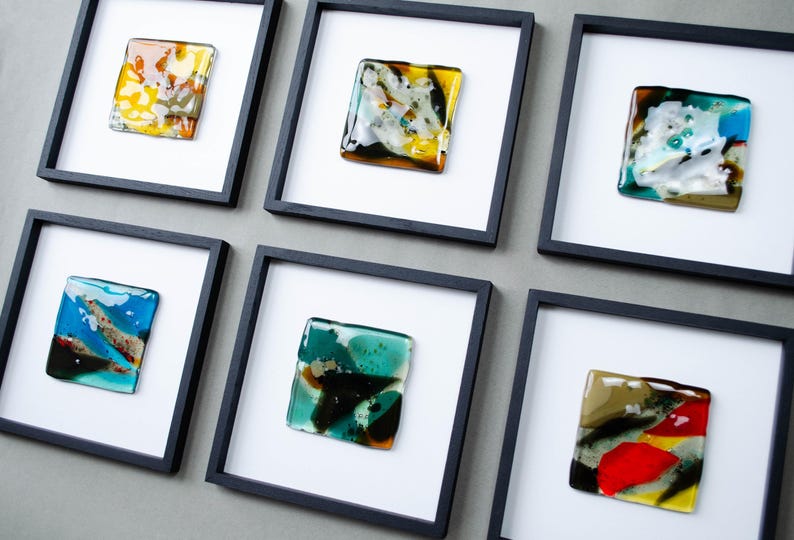May include: Six square black frames containing individual fused glass art pieces. Each piece features a unique abstract composition of vibrant colors including yellow, blue, green, red, and white, set against a plain white matting background.