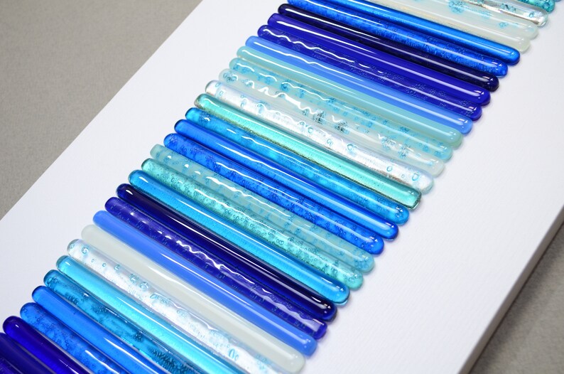 Pu&ograve; includere: Un'opera d'arte da parete realizzata con bacchette di vetro in varie tonalit&agrave; di blu, bianco e acquamarina. Le bacchette sono disposte in un motivo curvo, creando un effetto sfumato.