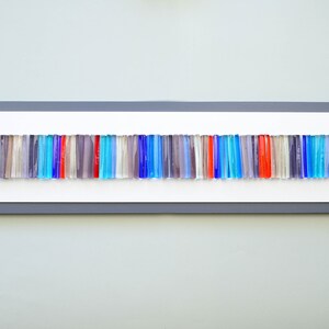 May include: A framed wall art piece featuring a horizontal row of colorful glass rods in shades of blue, red, and gray. The rods are arranged in a random pattern, creating a modern and abstract design.