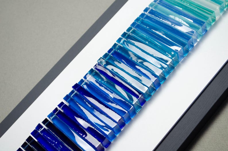 May include: A wall art piece made of glass tiles in shades of blue, teal, and green. The tiles are arranged in a gradient pattern, creating a wave-like effect.