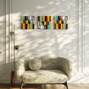 May include: Three square abstract artworks with a colourful, geometric design hang on a white wall. The art features horizontal blocks of colour including yellow, red, black, and blue. A beige sofa with a round cushion sits below the art.