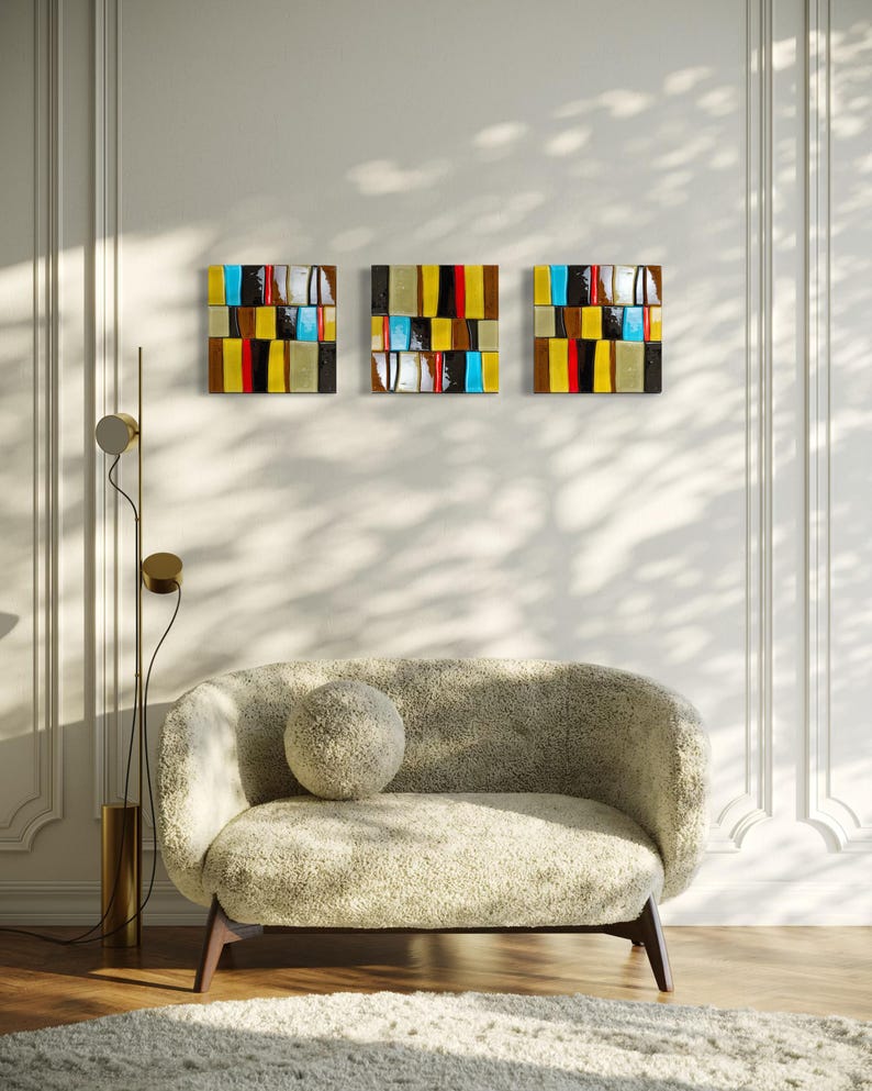 May include: Three square abstract artworks with a colourful, geometric design hang on a white wall. The art features horizontal blocks of colour including yellow, red, black, and blue. A beige sofa with a round cushion sits below the art.