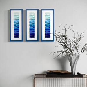 May include: Three framed pieces of abstract art with a gradient of blue shades. The frames are a dark blue color and the artwork is made of glass.