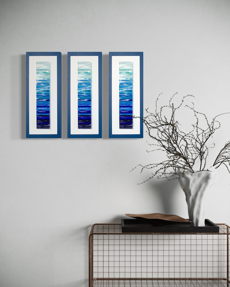 May include: Three framed pieces of abstract art with a gradient of blue shades. The frames are a dark blue color and the artwork is made of glass.