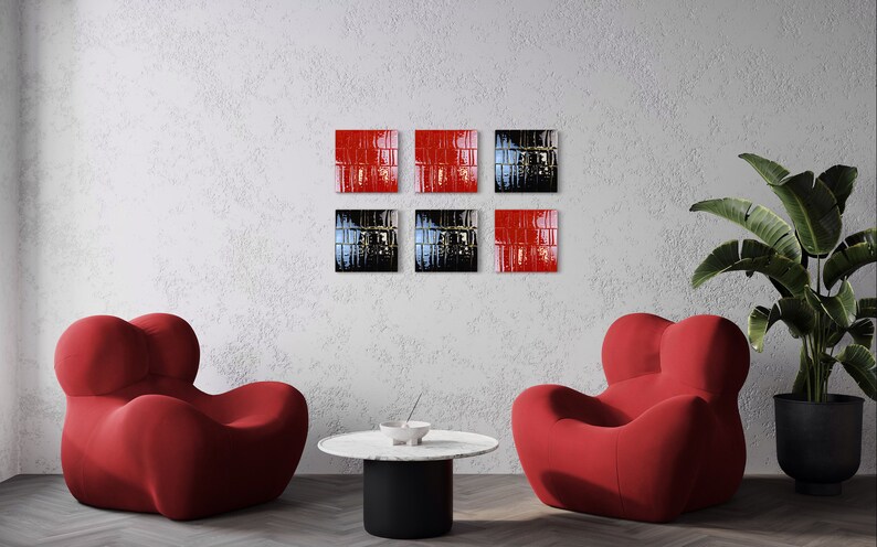 May include: Six square wall art pieces in red and black are displayed on a textured white wall. Two red armchairs and a round table with a black base are in front of the wall. A potted plant is on the right.