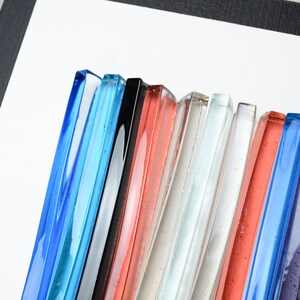 Pu&ograve; includere: Una collezione di piastrelle di vetro in vari colori, tra cui blu, nero, rosso, bianco e viola. Le piastrelle sono disposte in una fila, con i bordi di ogni piastrella visibili.
