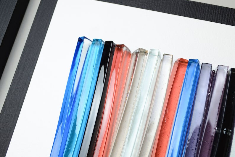 Pu&ograve; includere: Una collezione di piastrelle di vetro in vari colori, tra cui blu, nero, rosso, bianco e viola. Le piastrelle sono disposte in una fila, con i bordi di ogni piastrella visibili.
