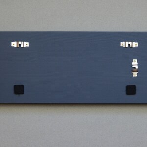 May include: The back of a dark gray rectangular wooden sign with two black square hanging hardware pieces and two silver hanging hooks.