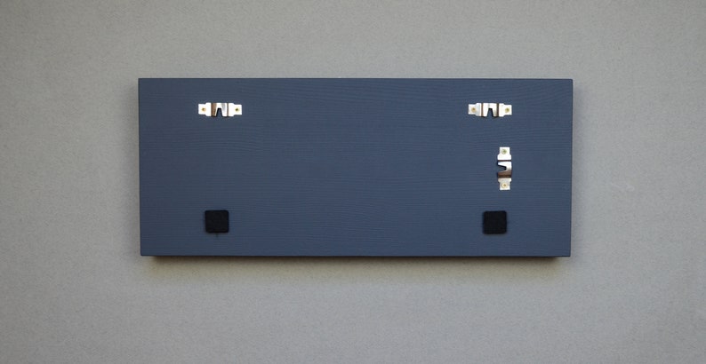 May include: The back of a dark gray rectangular wooden sign with two black square hanging hardware pieces and two silver hanging hooks.