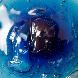 May include: A circular glass plate featuring a vibrant gradient of blue hues, ranging from deep navy at the center to a lighter turquoise at the edges. The surface is embedded with numerous small, clear air bubbles of varying sizes throughout.