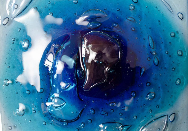 May include: A circular glass plate featuring a vibrant gradient of blue hues, ranging from deep navy at the center to a lighter turquoise at the edges. The surface is embedded with numerous small, clear air bubbles of varying sizes throughout.