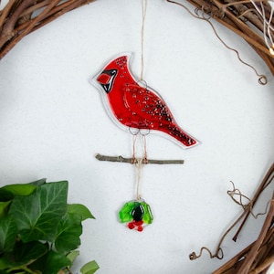 May include: A red cardinal bird ornament with a clear outline, perched on a small branch. The bird hangs from a string, with a small wreath below. The ornament is set against a brown wreath and white background.