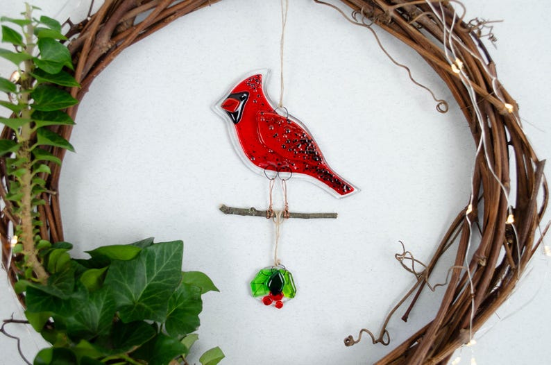 May include: A red cardinal bird ornament with a clear outline, perched on a small branch. The bird hangs from a string, with a small wreath below. The ornament is set against a brown wreath and white background.