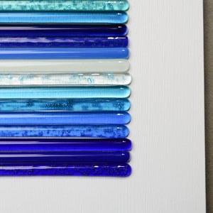 Pu&ograve; includere: Un'opera d'arte da parete realizzata con bacchette di vetro in tonalit&agrave; di blu, turchese e bianco. Le bacchette sono disposte in strisce orizzontali, creando un effetto sfumato.