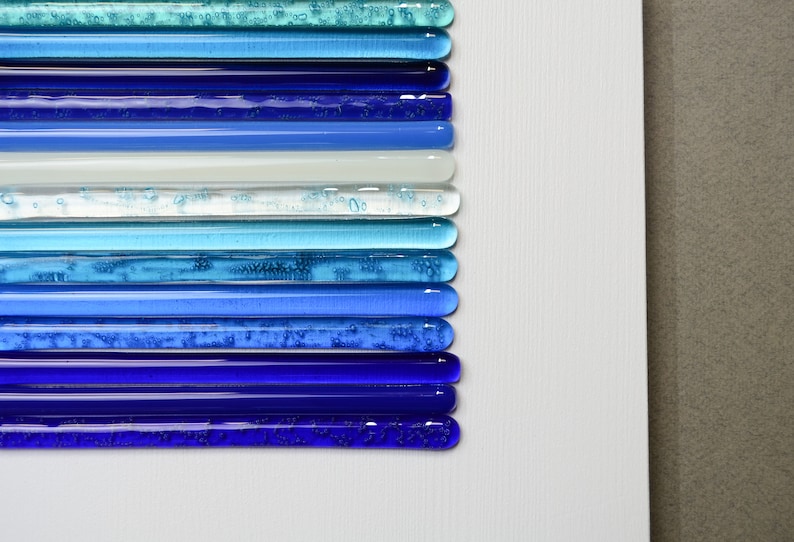 Pu&ograve; includere: Un'opera d'arte da parete realizzata con bacchette di vetro in tonalit&agrave; di blu, turchese e bianco. Le bacchette sono disposte in strisce orizzontali, creando un effetto sfumato.