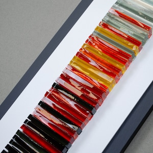 May include: A framed wall art piece featuring a vertical arrangement of colorful glass tiles. The tiles are arranged in a repeating pattern of black, red, yellow, and clear glass. The piece is mounted on a white background with a black frame.