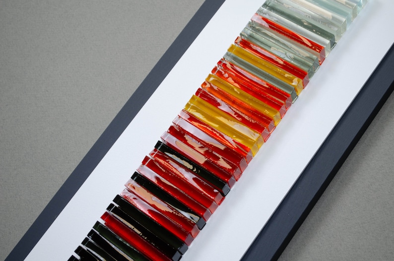 May include: A framed wall art piece featuring a vertical arrangement of colorful glass tiles. The tiles are arranged in a repeating pattern of black, red, yellow, and clear glass. The piece is mounted on a white background with a black frame.