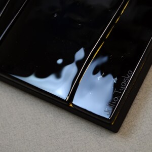 May include: Close-up of a rectangular black tray with a glossy finish. The tray has a series of parallel grooves and a small inscription that reads "Julia Tyasko". The tray is on a light grey surface.