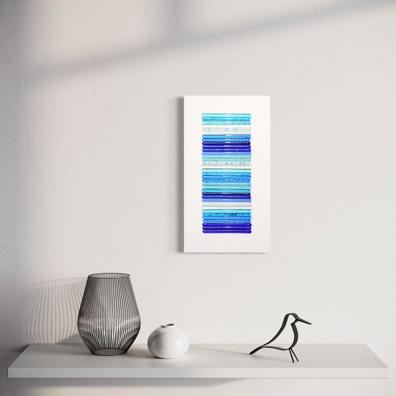 Pu&ograve; includere: Arte murale astratto con una disposizione verticale di pezzi di vetro blu, blu chiaro e bianco. I pezzi di vetro sono impilati uno sopra l'altro, creando un effetto a strati.