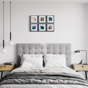 May include: A bedroom interior featuring a gray tufted headboard, a bed with white bedding, two light wood nightstands with black legs, two hanging globe pendant lights, a black desk lamp, and a gallery wall of six square framed abstract art prints.
