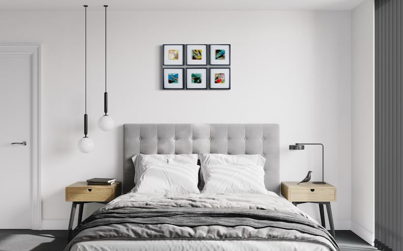 May include: A bedroom interior featuring a gray tufted headboard, a bed with white bedding, two light wood nightstands with black legs, two hanging globe pendant lights, a black desk lamp, and a gallery wall of six square framed abstract art prints.