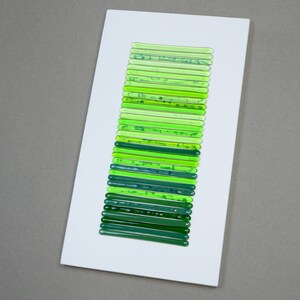 May include: A white rectangular frame with a vertical arrangement of green glass rods of varying shades, creating a gradient effect.