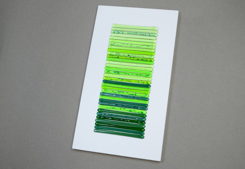 May include: A white rectangular frame with a vertical arrangement of green glass rods of varying shades, creating a gradient effect.