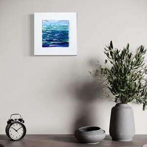 May include: Abstract art piece featuring a textured, layered design in shades of blue and green, mounted on a white square. The artwork evokes a sense of water or landscape. The piece is displayed on a neutral-toned wall.