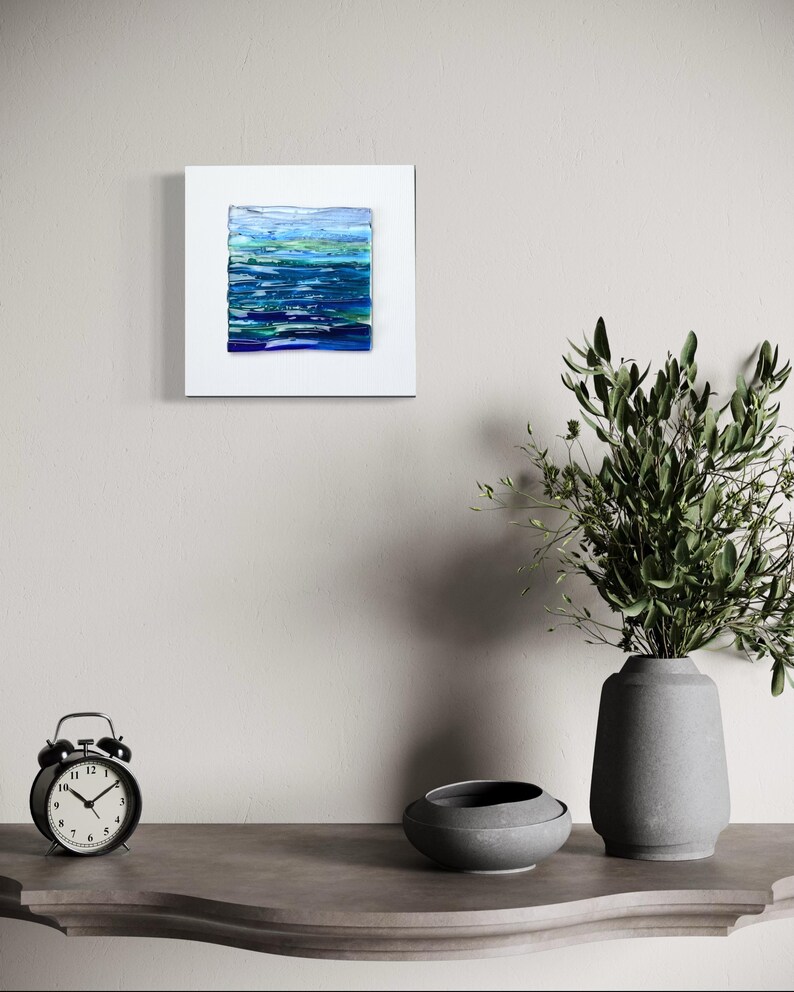 May include: Abstract art piece featuring a textured, layered design in shades of blue and green, mounted on a white square. The artwork evokes a sense of water or landscape. The piece is displayed on a neutral-toned wall.